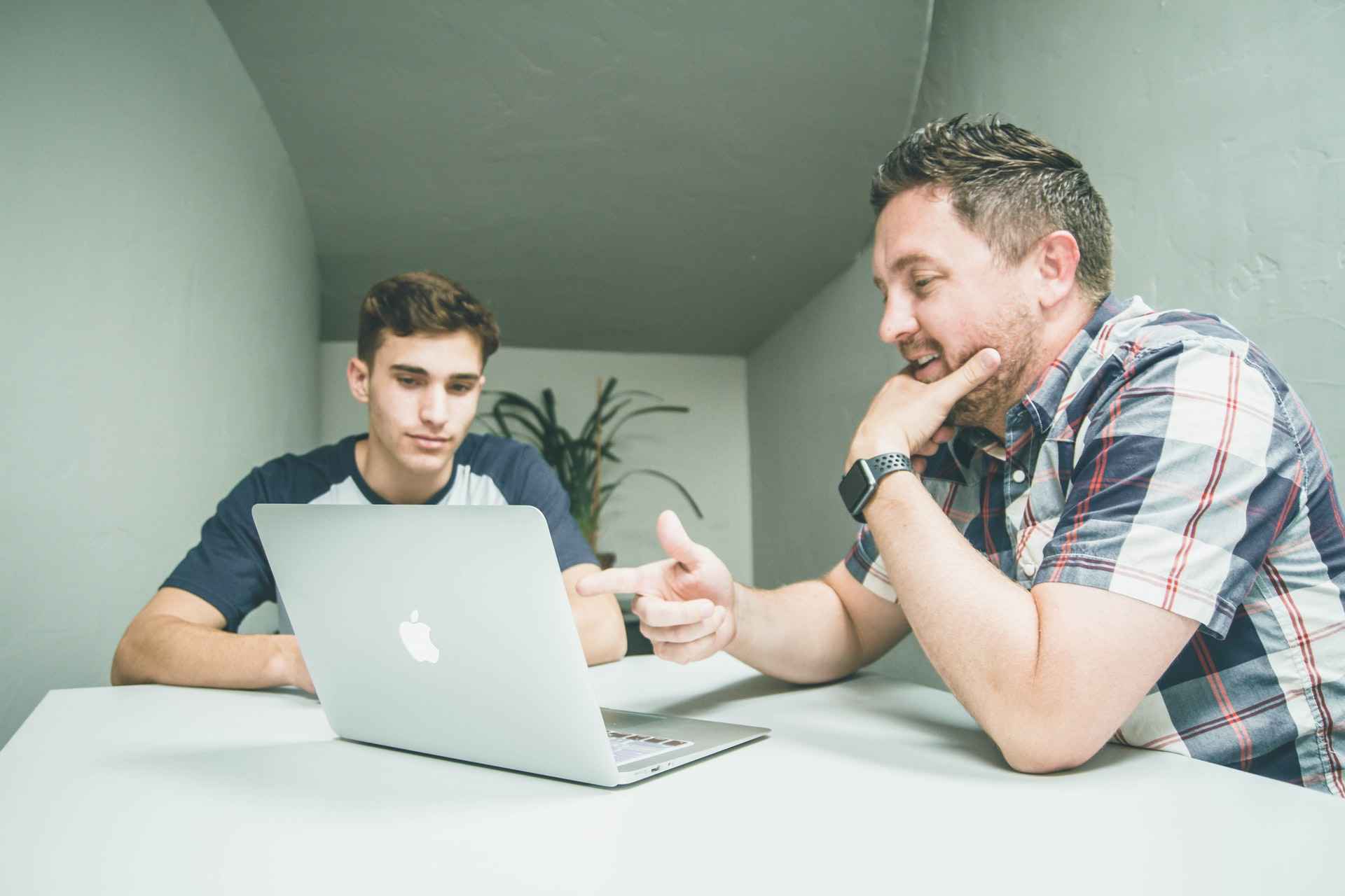 Two men sitting at a desk looking at a laptop, one man pointing to it while giving the other different feedback examples